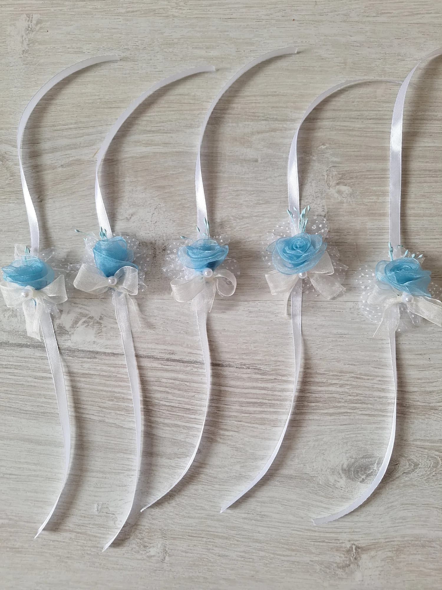 Five wrist corsages, each with a pale blue organza rose, an ivory organza bow and a pearl, tied to a white satin ribbon.